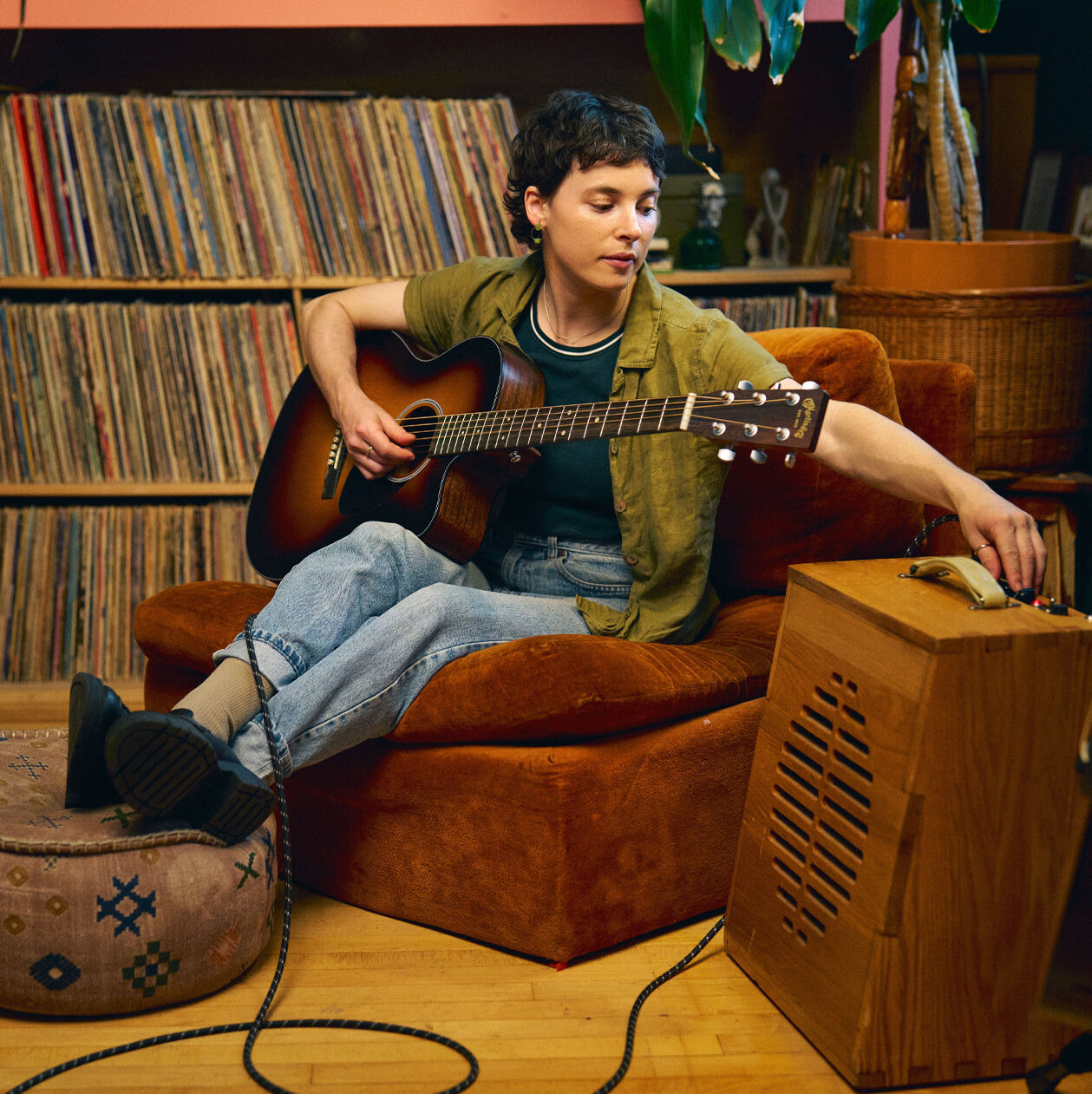 A person sitting on a lounge chair with records behind them playing an acoustic guitar