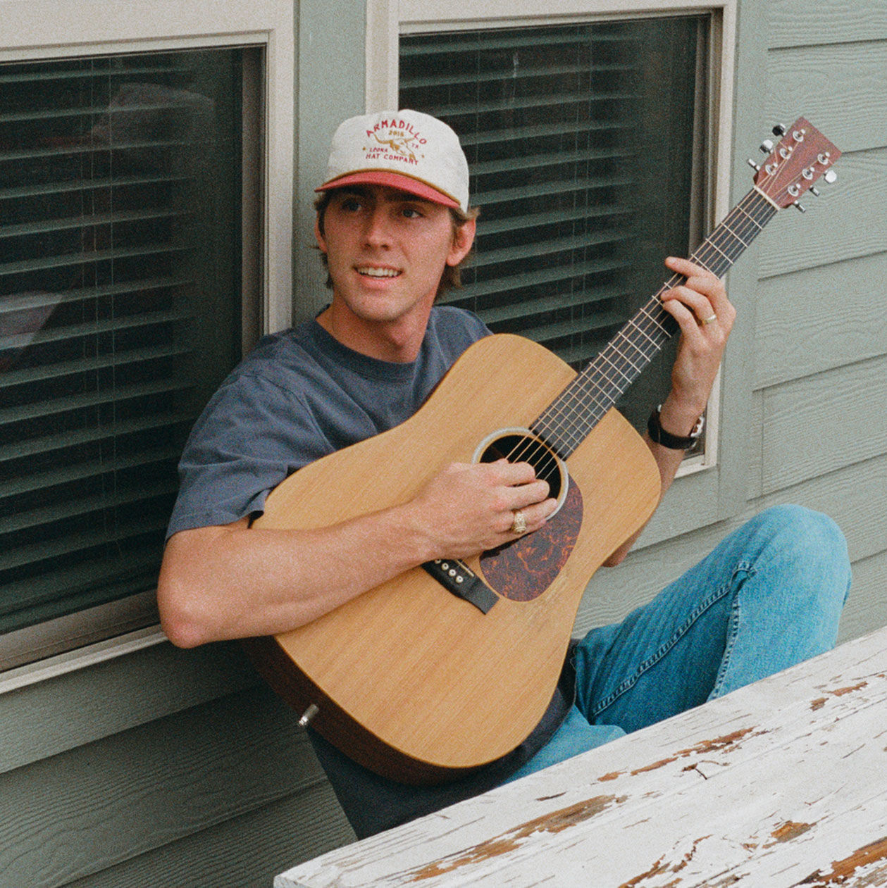 Photo of Dylan Gossett sitting on a porch playing an acoustic guitar in a ballcap.