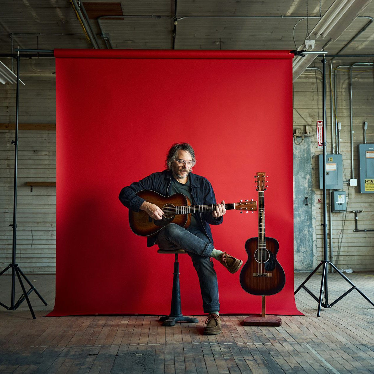 Jeff Tweedy in front of a red backdrop with his acoustic guitars
