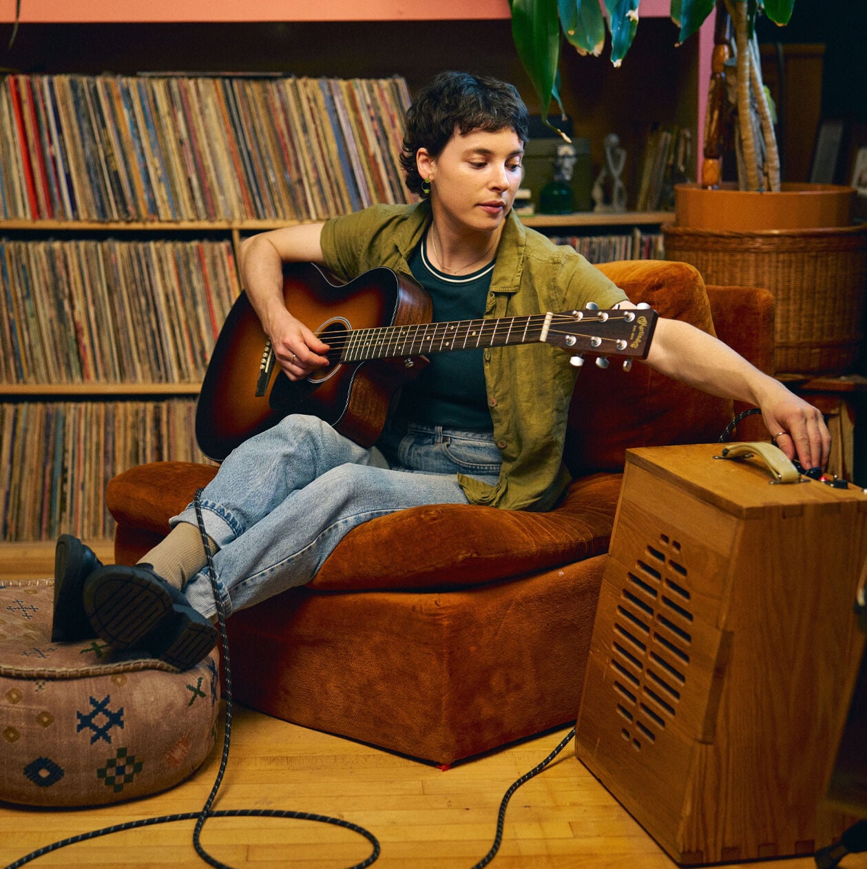 A person sitting on a lounge chair with records behind them playing an acoustic guitar