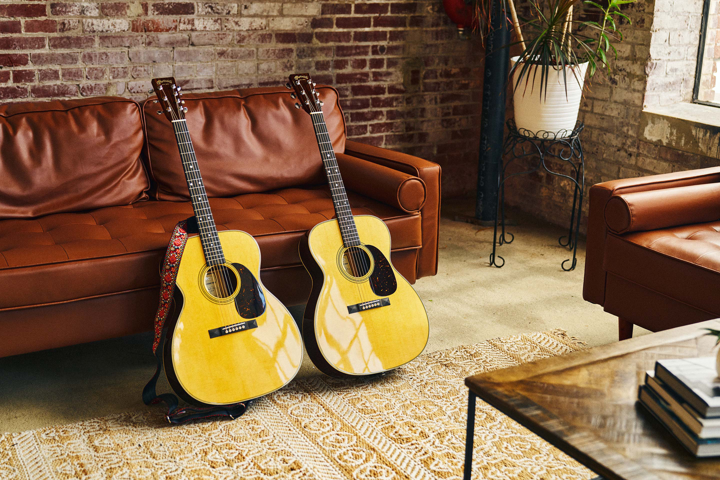 Two guitars sitting side by side leaning against a brown leather couch in a brick room